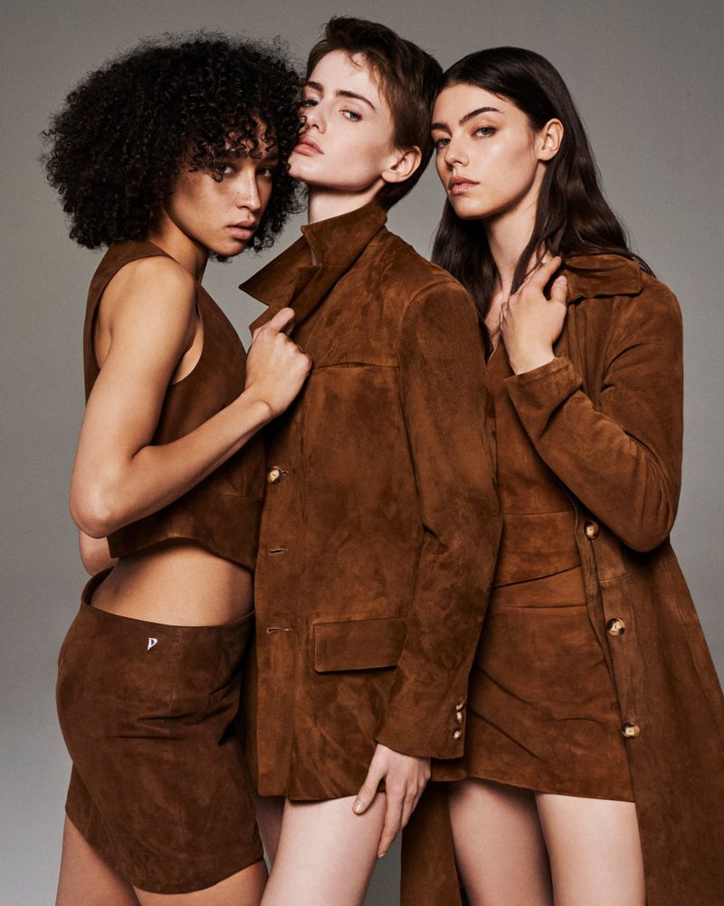 Three fashion models wearing stylish brown suede outfits pose in a studio with a neutral grey backdrop, showcasing modern style.