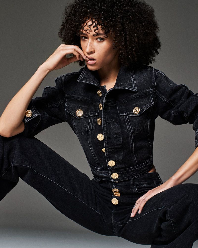Model with short curly hair posing in a cropped black denim jacket and matching jeans with large gold buttons against a grey studio backdrop.