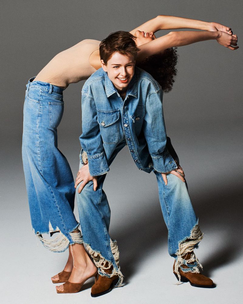 Two models posing playfully in distressed blue denim outfits and brown footwear against a seamless grey studio background.