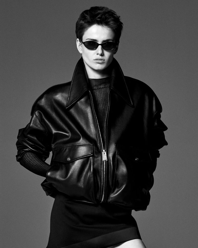 Model wearing a black oversized leather jacket, dark sunglasses, and black mini skirt against a grey studio backdrop.