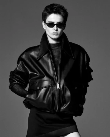 Model wearing a black oversized leather jacket, dark sunglasses, and black mini skirt against a grey studio backdrop.