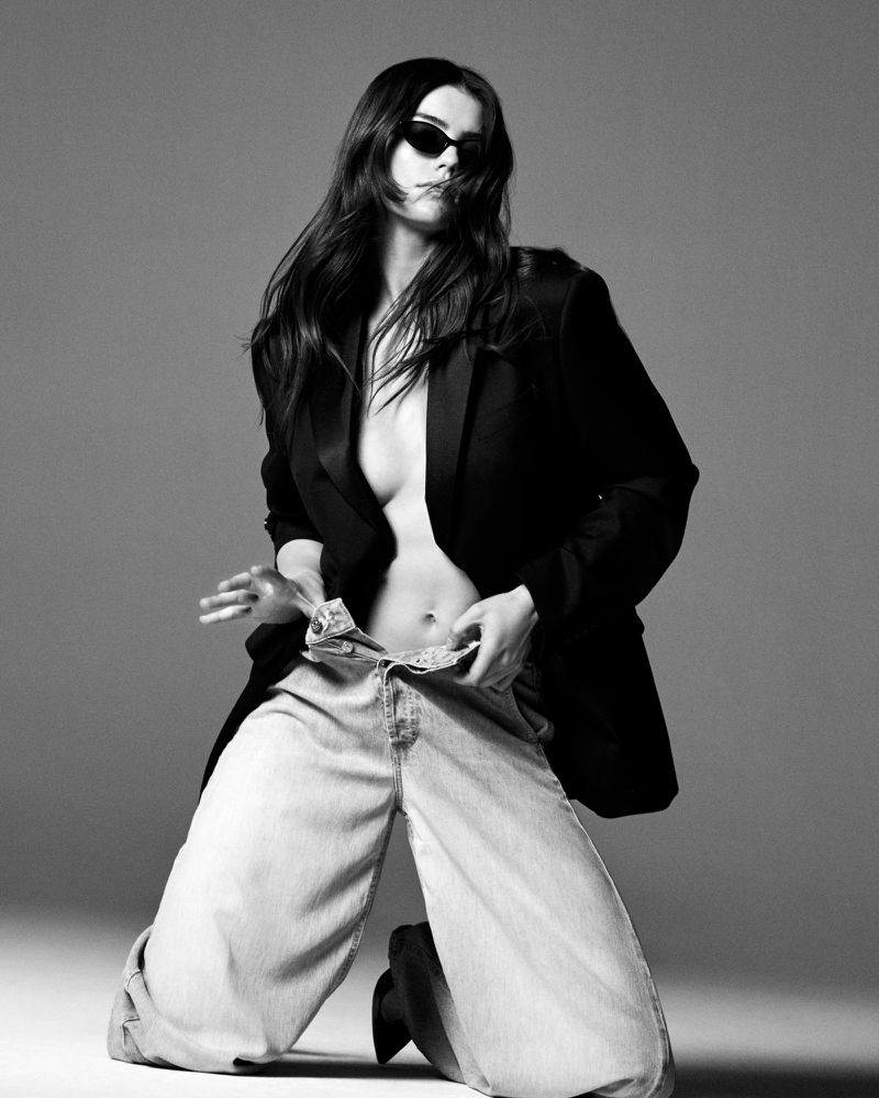 Model kneeling in an open black blazer and light wash wide-leg jeans with sunglasses against a grey studio backdrop.