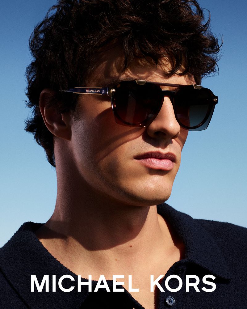 Model Francesco Ruggiero wearing dark sunglasses and a navy polo against a clear blue sky in a Michael Kors eyewear campaign.