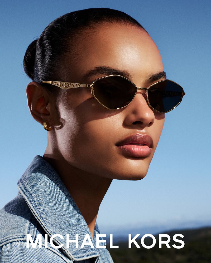 Model wearing geometric gold-framed sunglasses and a denim jacket against a bright blue sky for Michael Kors.