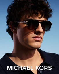 Model Francesco Ruggiero wearing dark sunglasses and a navy polo against a clear blue sky in a Michael Kors eyewear campaign.