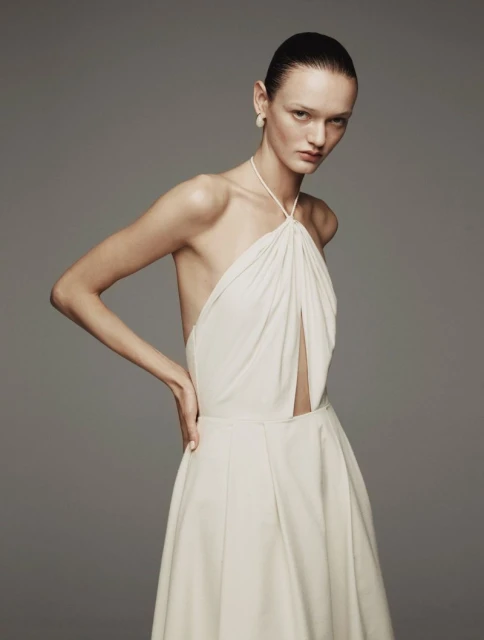 Fashion portrait of a female model with slicked-back hair wearing a white halter dress against a neutral grey background in a photo studio.