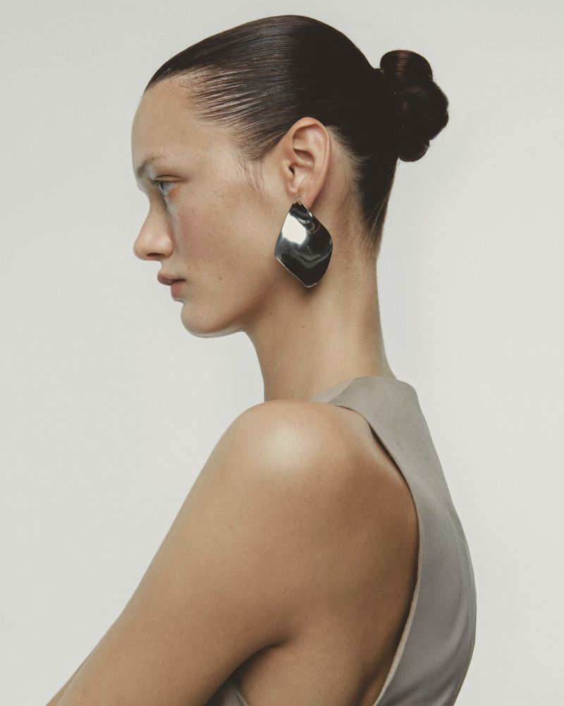 Profile of a woman with a low bun and large silver earring against a plain backdrop in a fashion shot with neutral color grading.