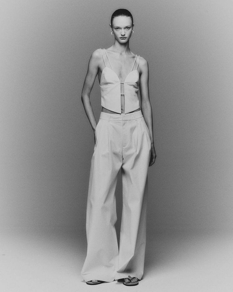 A woman in a light-colored matching top and wide-leg pants stands in a photo studio, hands in her pockets, in a black-and-white fashion shot.