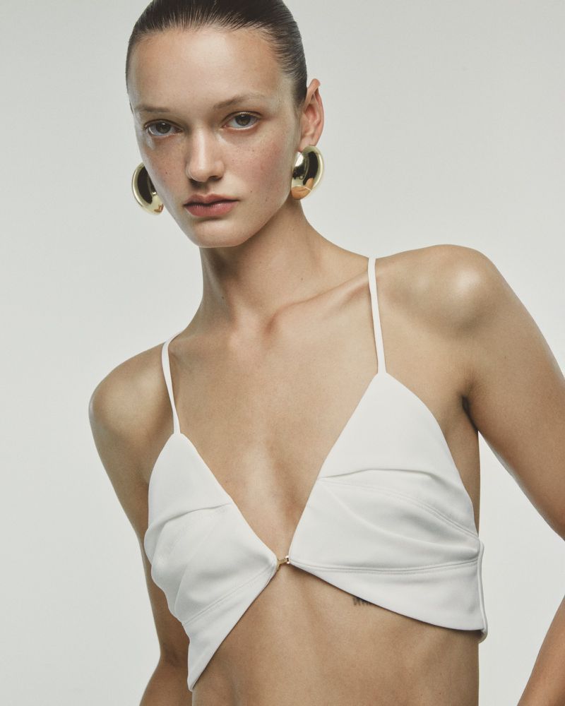 Close-up studio shot of a woman wearing a white bra top and large gold hoop earrings against a neutral backdrop, fashion studio.
