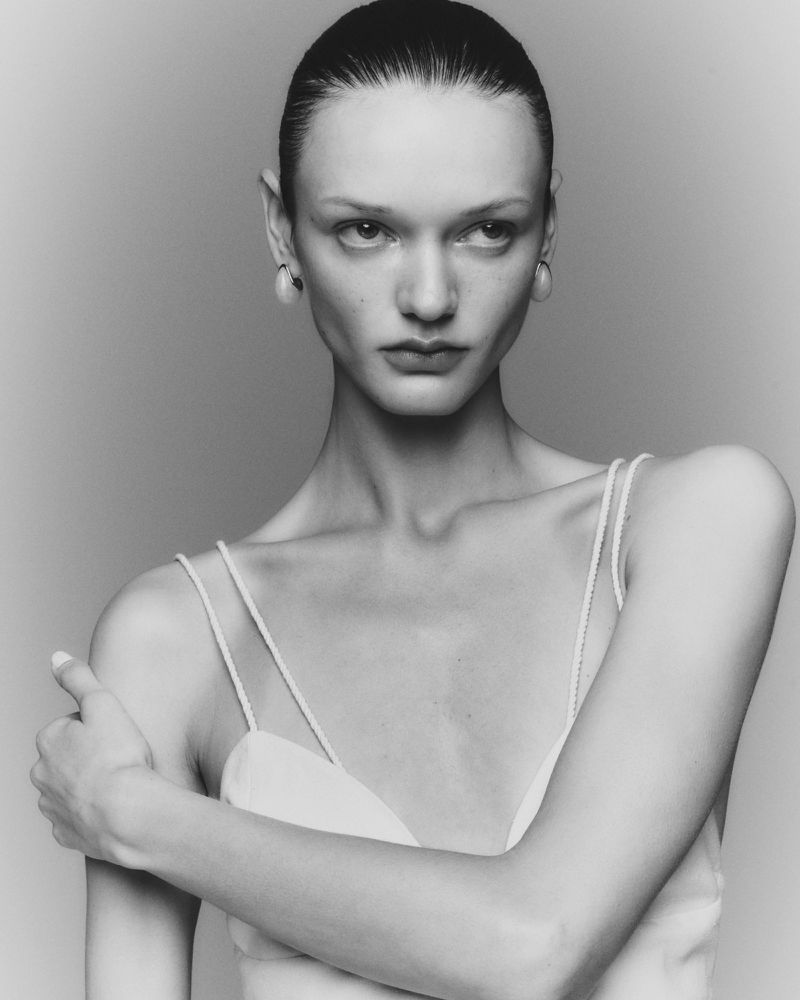 Monochrome portrait of a fair-skinned woman with slicked-back hair in a light-colored top, her arms crossed, against a neutral studio backdrop.