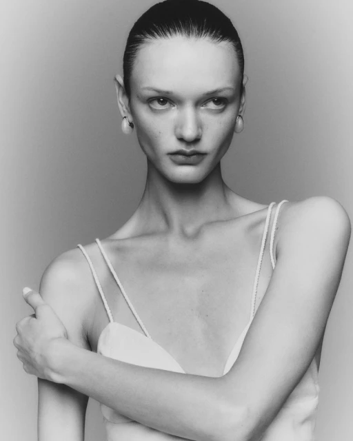 Monochrome portrait of a fair-skinned woman with slicked-back hair in a light-colored top, her arms crossed, against a neutral studio backdrop.