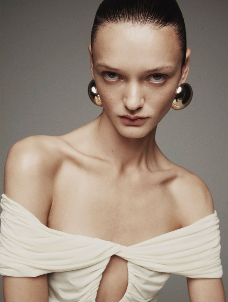 Close-up of a woman with slicked-back hair wearing large, round gold earrings and a cream-colored dress with a keyhole cutout at the chest.