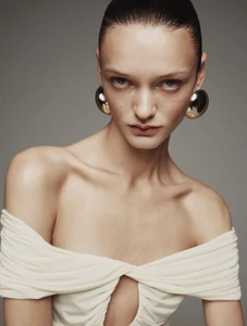 Ali Honcharuk wearing large gold dome earrings and an off-the-shoulder draped white top against a grey studio backdrop.