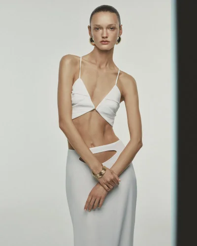 Model wearing a white two-piece crop top and matching skirt with cut-out details against a pale grey studio background.