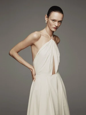 Model Ali Honcharuk posing with hands on hips wearing an ivory halter-neck dress against a plain grey studio backdrop.