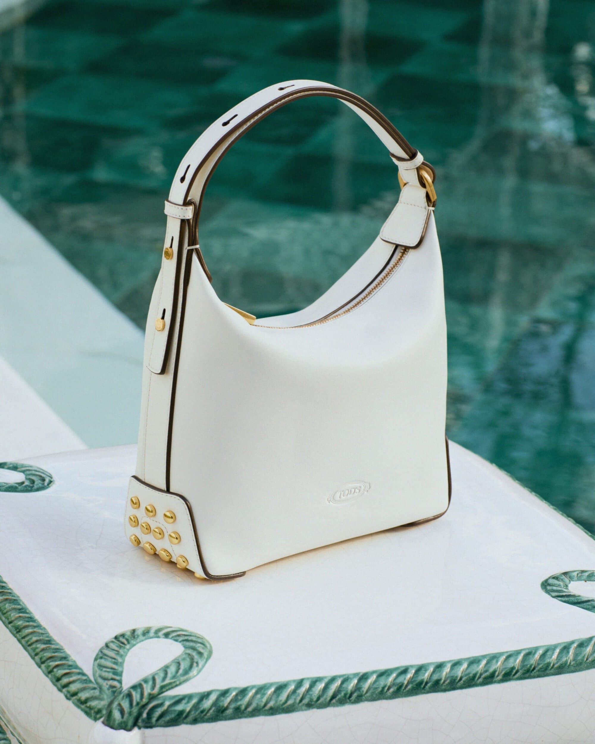 White leather Tod's hobo handbag with gold stud hardware resting beside a turquoise tiled swimming pool.