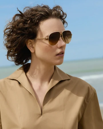 Woman with curly brown hair wearing aviator sunglasses and a beige collared shirt against a coastal sky and sea background.