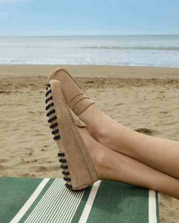 Crossed feet wearing beige suede driving loafers resting on a green striped deckchair at a sandy beach overlooking the sea.