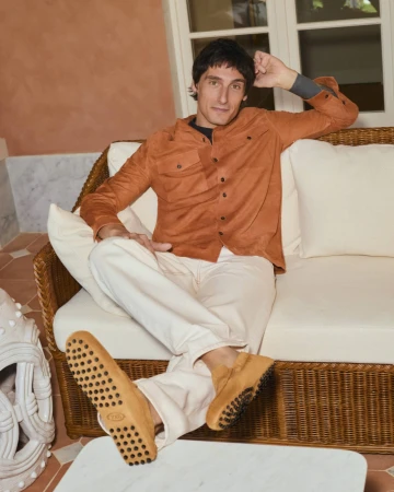 A man relaxing on a wicker sofa wearing a terracotta overshirt, off-white trousers, and tan suede driving loafers.