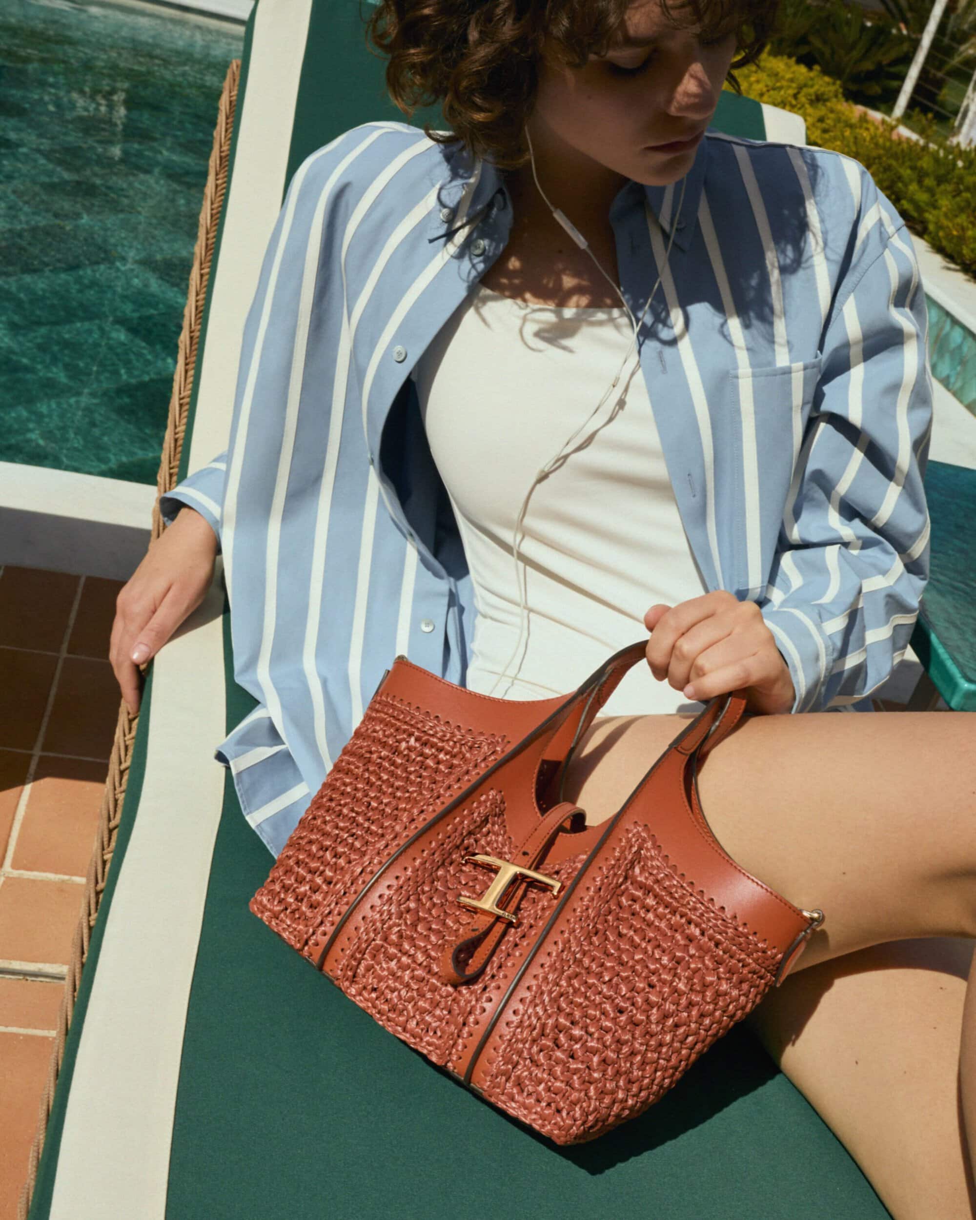 Model reclining on a poolside sun lounger holding a brown woven leather handbag with gold hardware.