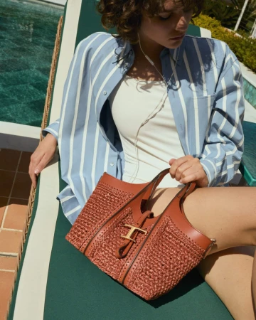 Model reclining on a poolside sun lounger holding a brown woven leather handbag with gold hardware.