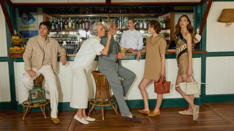 Group of people sitting and standing at a bar inside, wearing fashionable outfits and holding bags, with a bartender in the background.