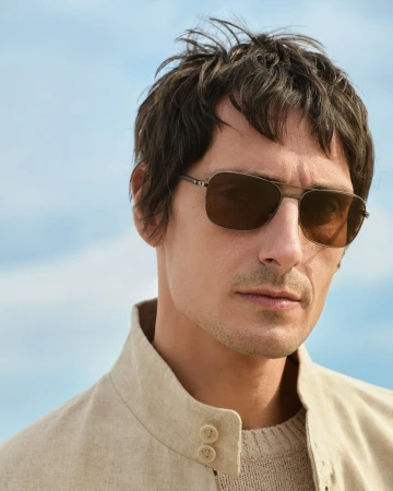 Close-up portrait of a man wearing dark aviator sunglasses and a beige linen high-collar jacket against a blue sky.