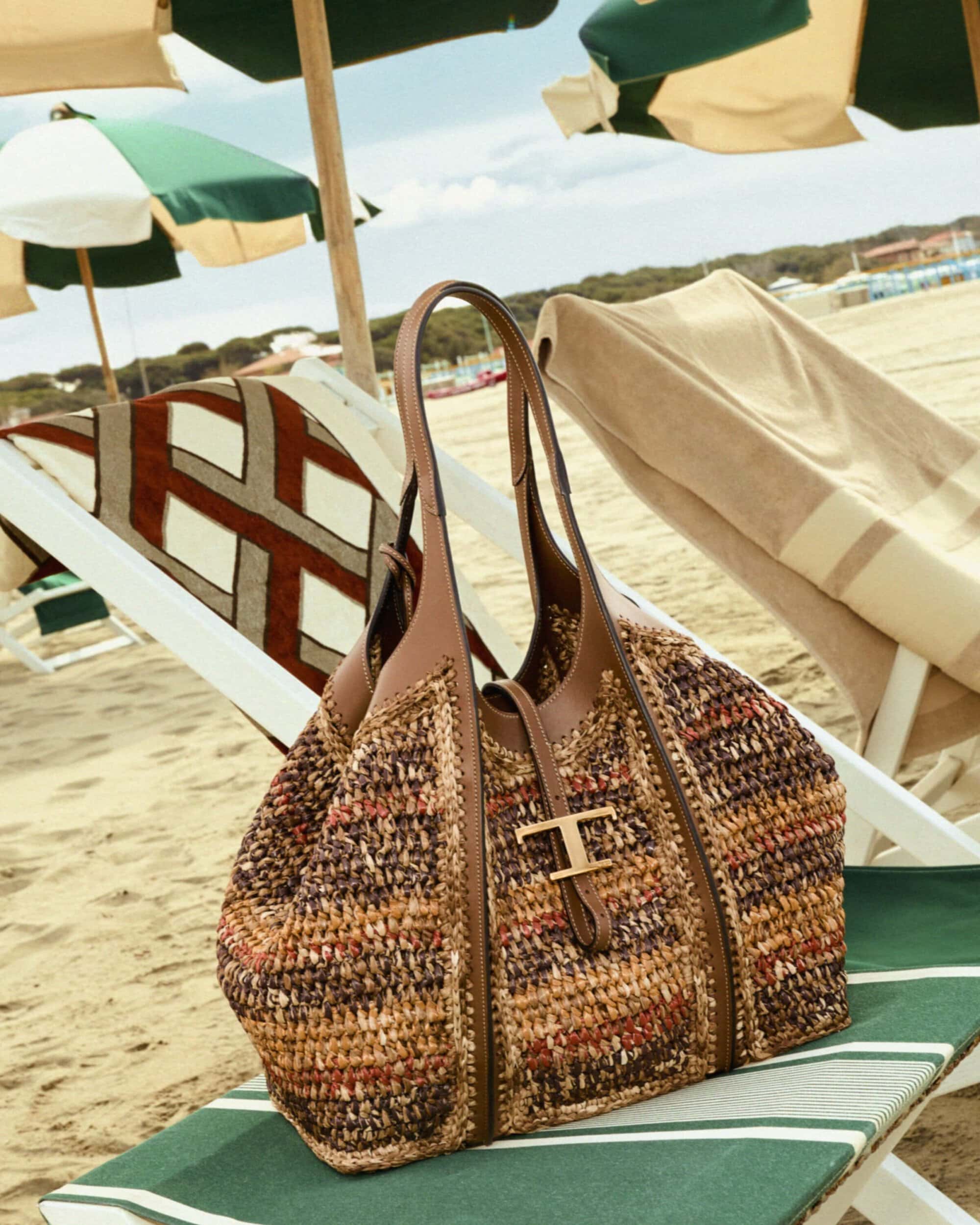 A woven earth-tone tote bag with brown leather handles sits on a green striped beach chair under sun umbrellas.