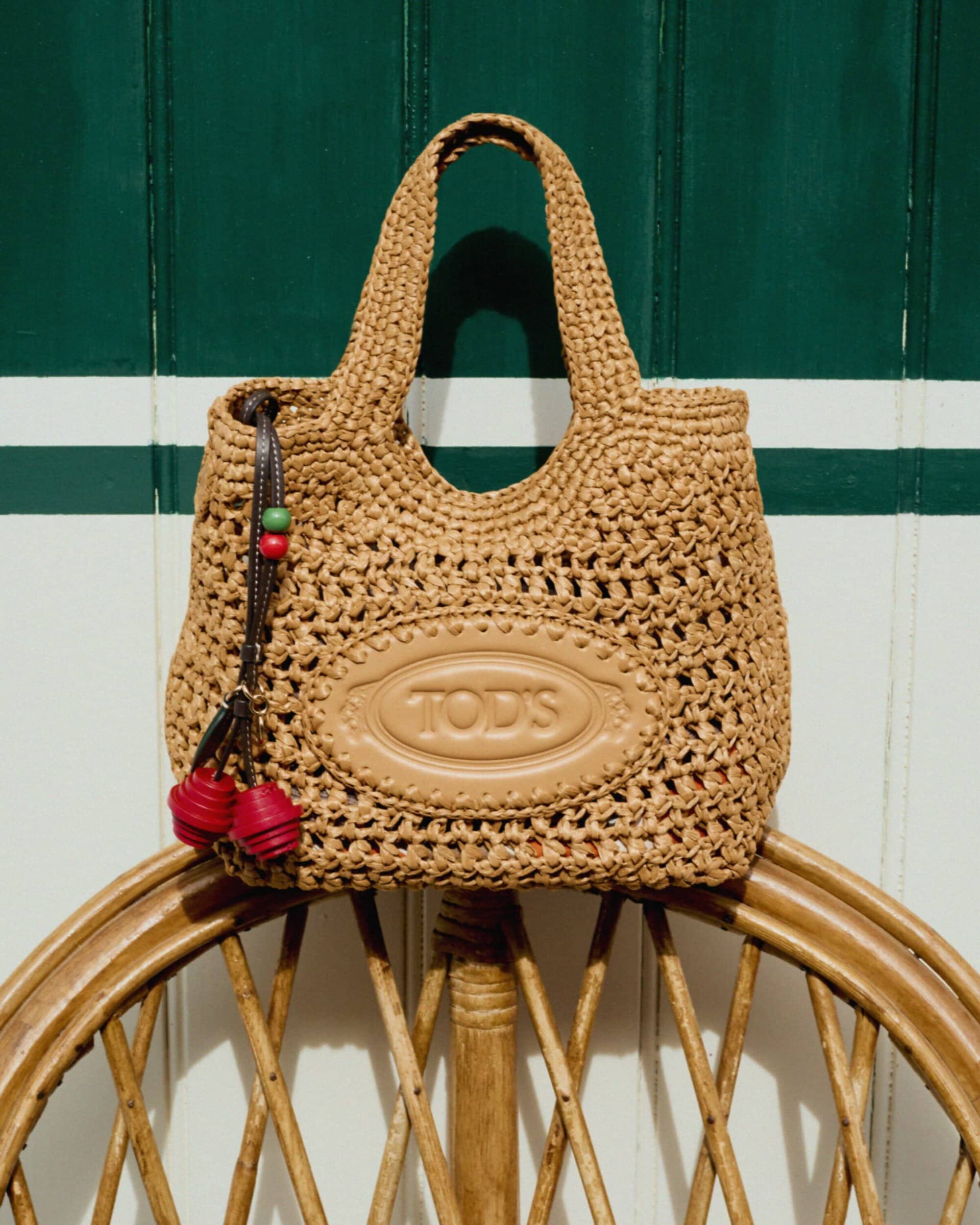Tod's woven raffia handbag with an embossed leather logo patch resting on a rattan chair against a green and white striped wall.