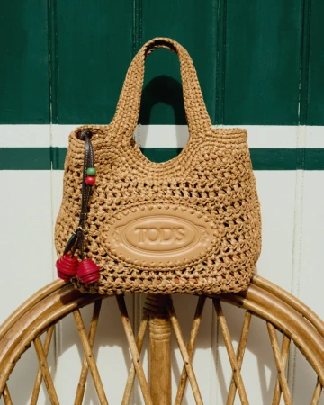 Tod's woven raffia handbag with an embossed leather logo patch resting on a rattan chair against a green and white striped wall.