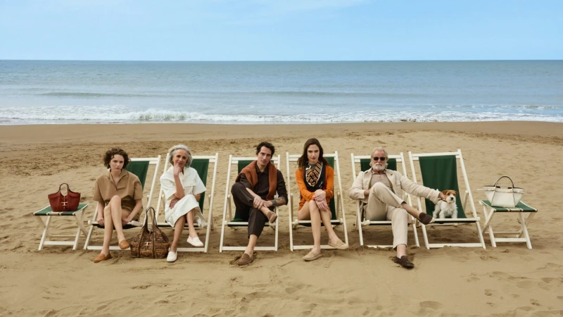 Five models and a small dog sit in green deckchairs on a sandy beach wearing neutral luxury apparel and loafers.