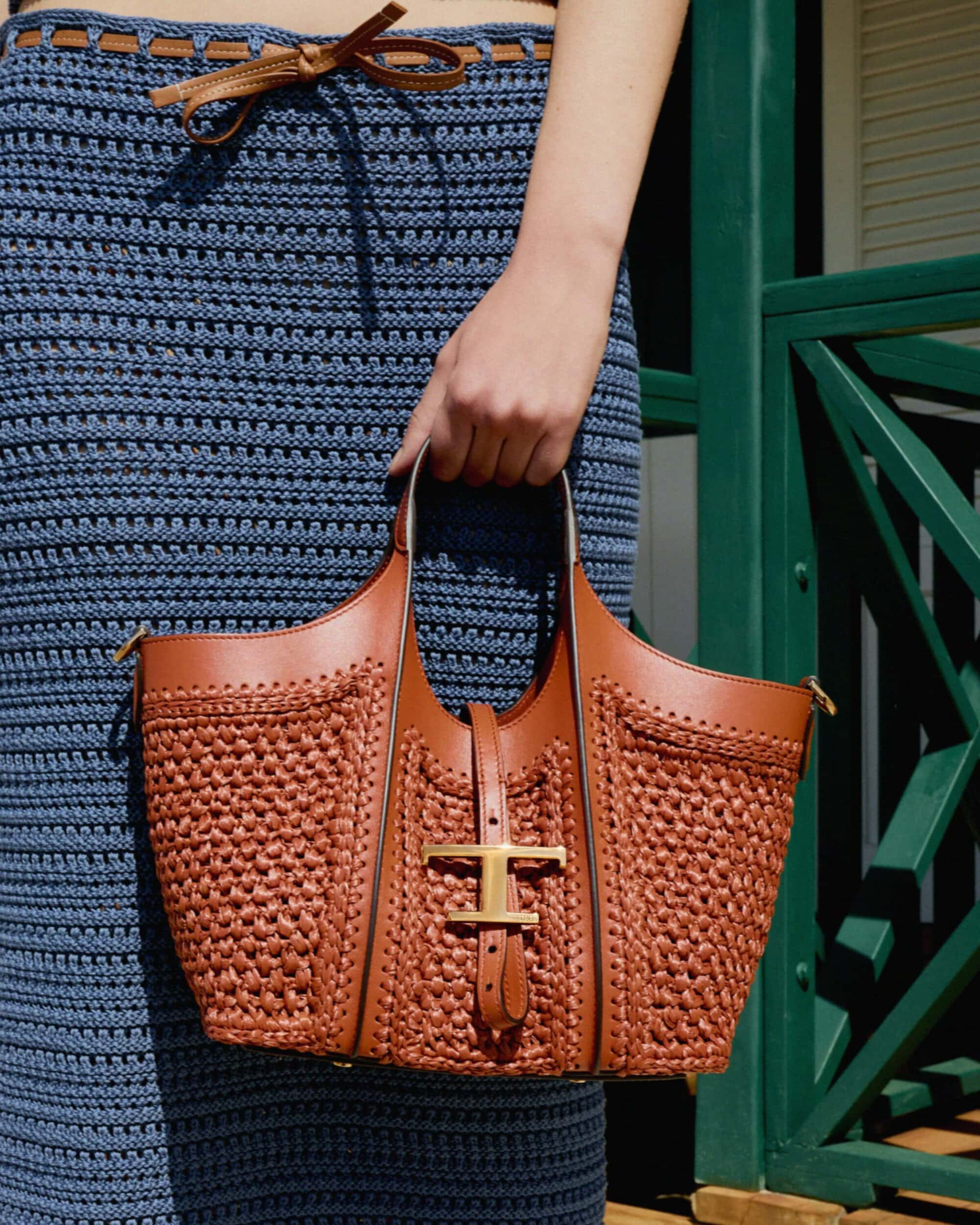 Close-up of a person wearing a blue crochet knit skirt holding a brown woven leather tote bag with a gold metal T logo.