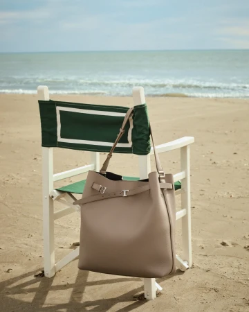 Taupe leather tote bag hanging on a white wooden director chair with green canvas on a sandy beach by the sea.