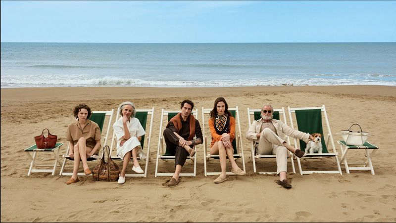 Group of six adults seated on green and white folding deck chairs on a sandy beach with the ocean in the background for Tod's campaign.