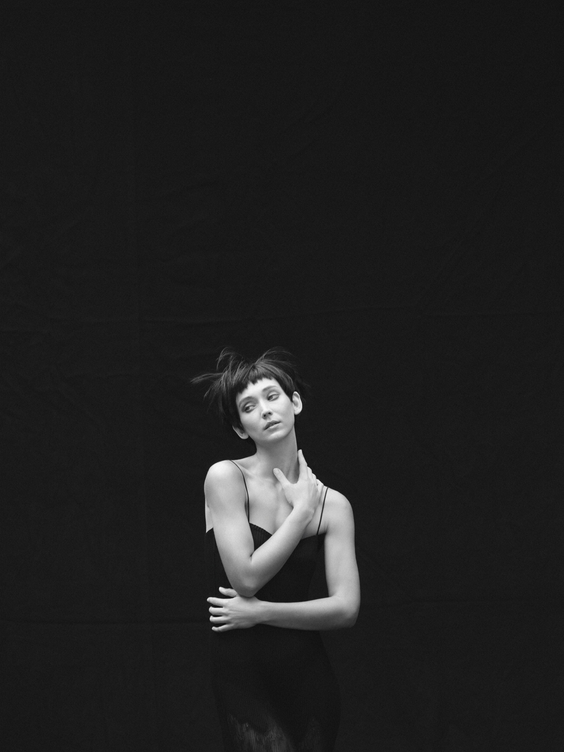 Monochrome portrait of a female model with short hair wearing a black dress, hand at neck, against a dark, minimalist background.
