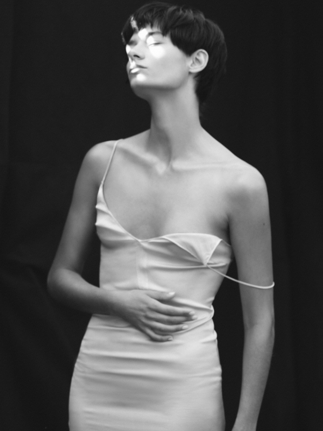 Monochrome portrait of a young woman with short dark hair wearing a light-colored dress against a dark background with dramatic lighting.