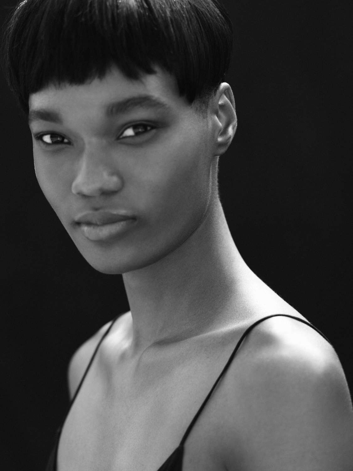 Monochrome portrait of a young Black woman with a short bowl cut, wearing a spaghetti strap top, set against a dark background.