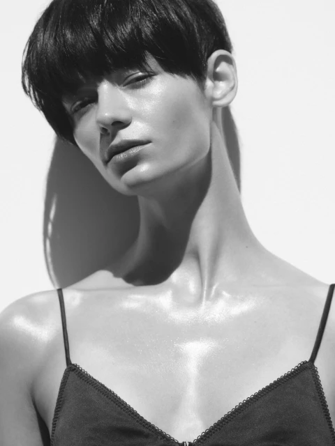 Close-up portrait of a young adult woman with short dark hair, wearing a black camisole in front of a bright white background.