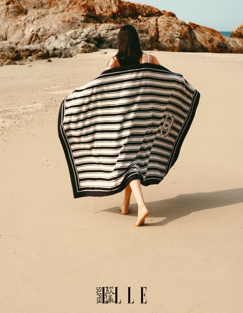 Model Jiahui Zhang walks barefoot along a sandy beach wearing a black and white striped Chanel blanket draped over her shoulders.
