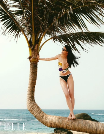Model Jiahui Zhang standing on a curved palm tree trunk overlooking the ocean, wearing sunglasses and swimwear.