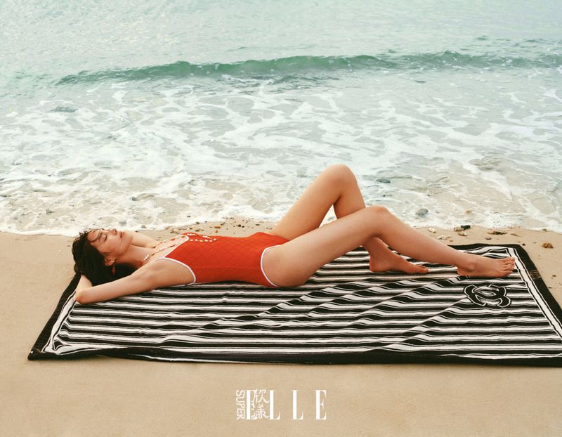 Model Jiahui Zhang lying on a striped beach towel wearing a red one-piece swimsuit on the sand near ocean waves.