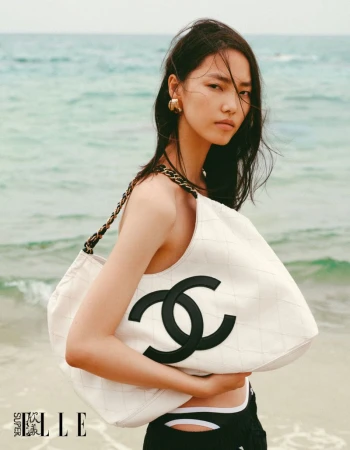Model Jiahui Zhang holding an oversized white quilted Chanel logo tote bag on a sandy ocean beach.