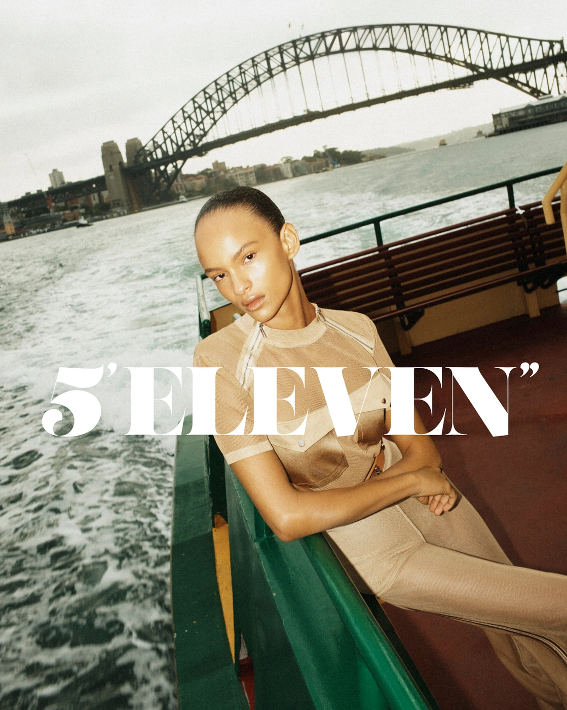 Model wearing a tan outfit leaning on a green boat railing with a large bridge and water visible in the background