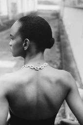 Black and white portrait from behind of a Black woman with a sleek low bun wearing a bold silver chain necklace and backless top.