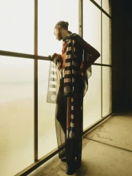Model standing by large gridded window wearing a patterned sheer scarf and dark garments for 5'Eleven The Islands Issue