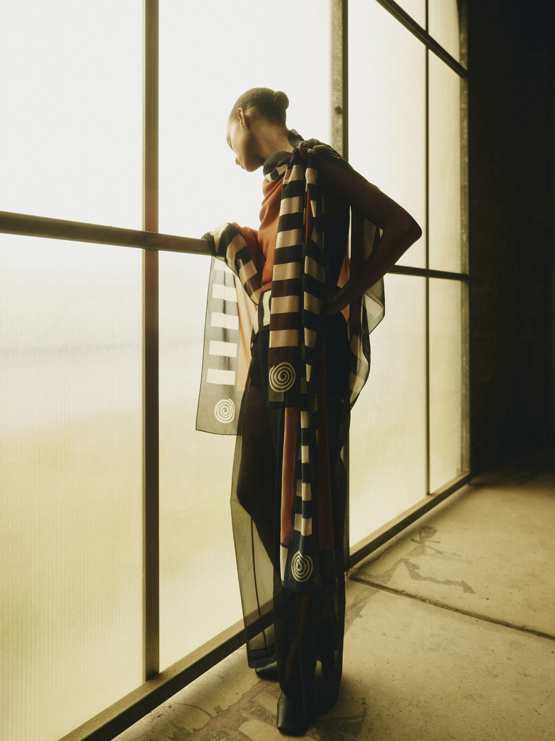 Woman in a patterned poncho leans against a window frame in an indoor industrial setting, looking out over a muted cityscape.