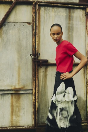 Woman in red top and skirt with a faded print stands against weathered metal door in an outdoor setting; fashion editorial.