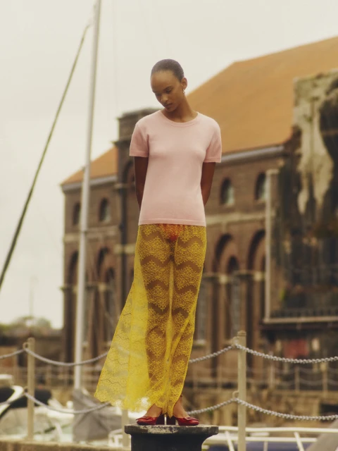 A woman stands outside, wearing a pink top and a long yellow lace skirt, in front of a brick building under a cloudy sky.