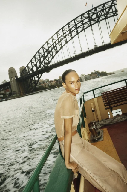 Fashion shot of a model in beige outfit posing on a boat with the Sydney Harbour Bridge in the background on an overcast day.