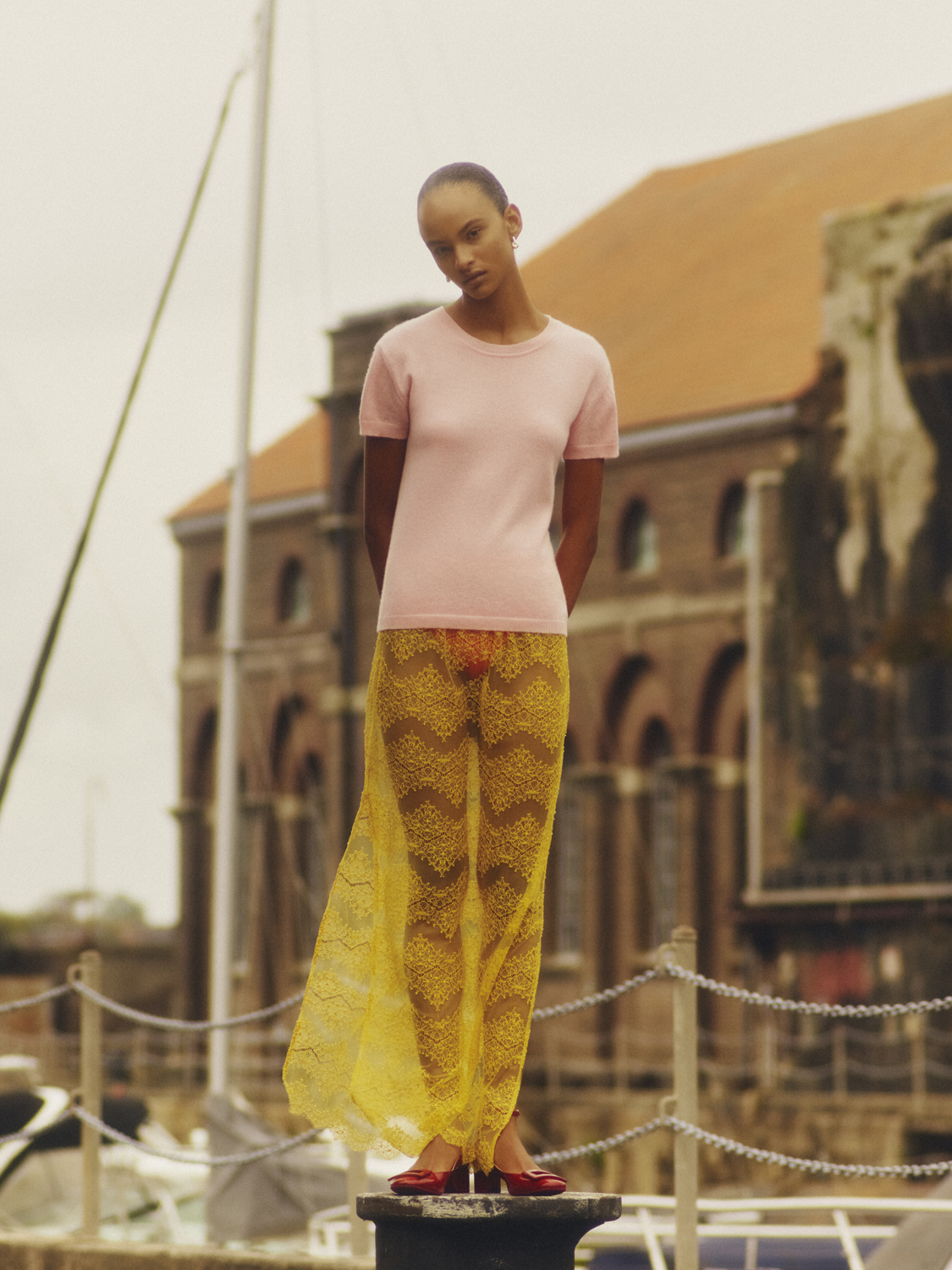 Fashion shot of a slender, light-skinned model in a pink top and yellow lace skirt posing on bollard with building in background.